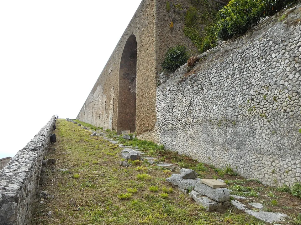 A grassy ramp with scattered squared stones climbing steeply between a low parapet on the left and a high wall of small rough stones on the right, towards a tall arch in a massive wall at the top.