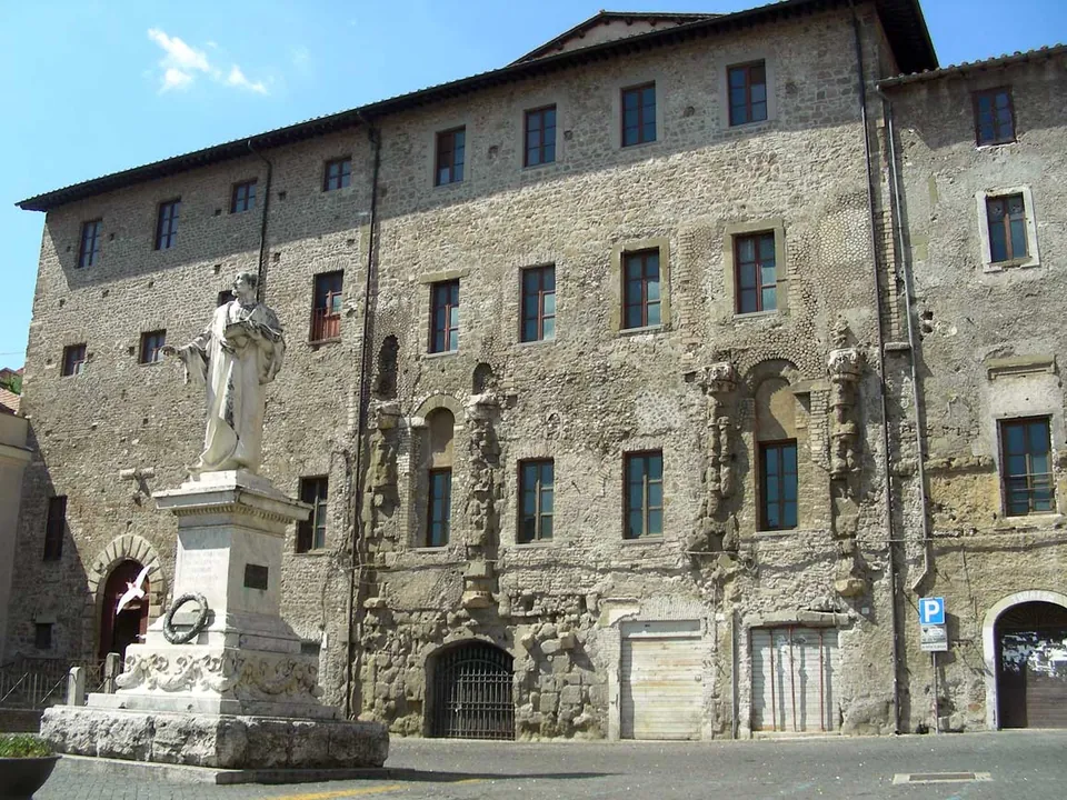 A tall stone palace front with rows of windows, the lower storeys studded with the worn stumps of ancient half-columns and carved fragments, and a white statue on a pedestal in front.