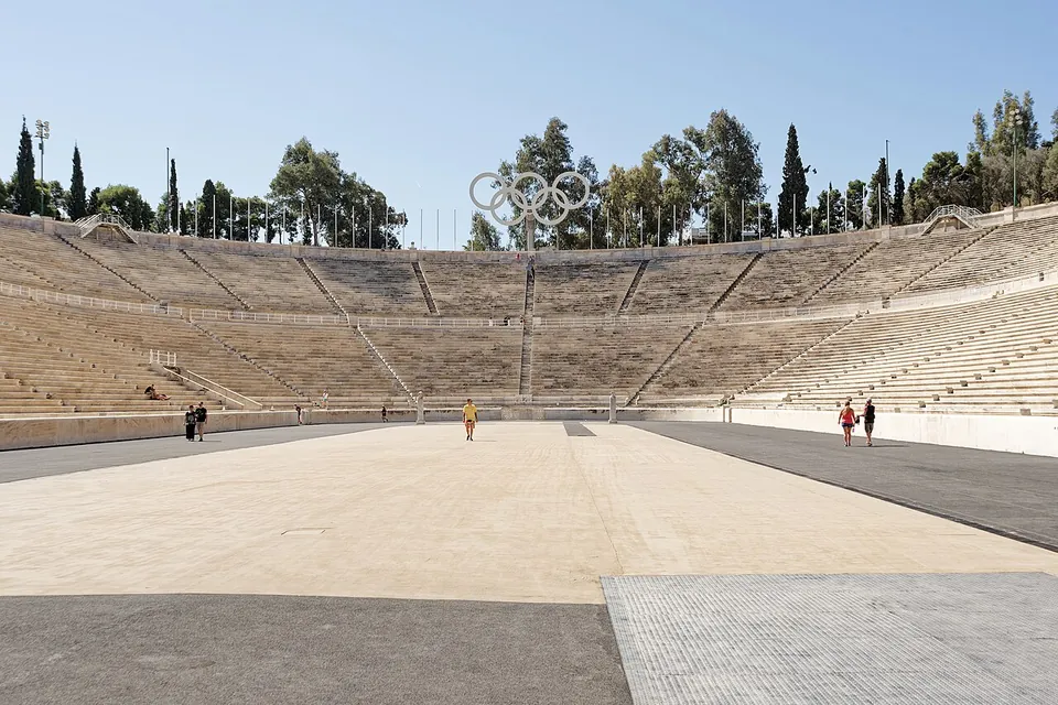 A long stadium of white marble seen from its open end, tiers of marble seats rising on both sides and round the curved far end, which carries a large sculpture of the Olympic rings, with a few people on the track.