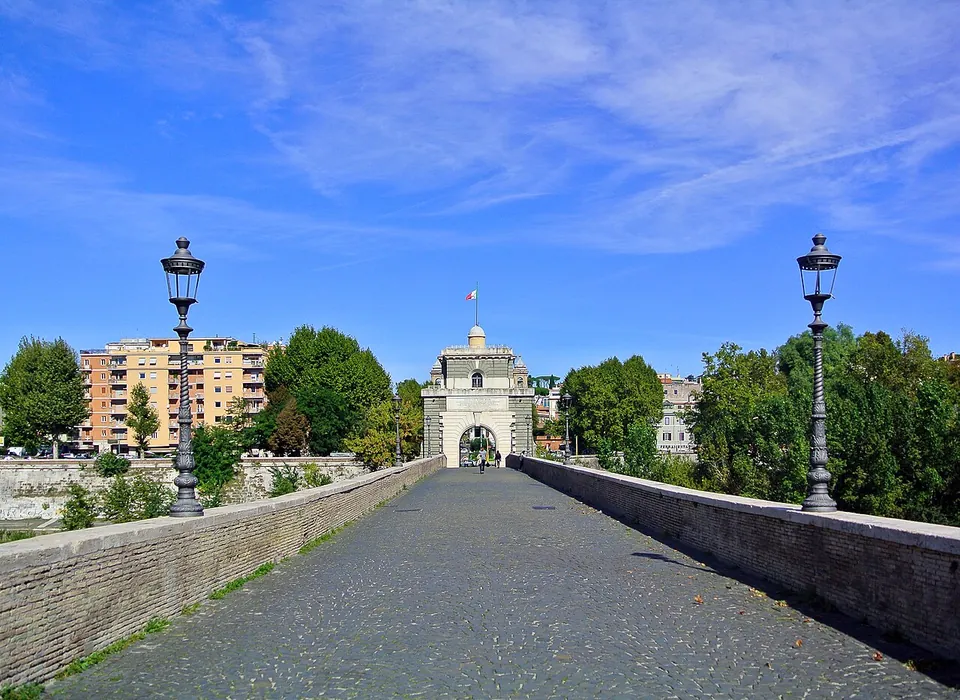 A cobbled walkway between low parapets with old-fashioned lamp posts, leading to a pale stone gate tower with an arched passage, under a blue sky.