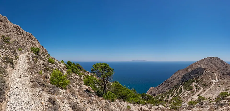 A wide view from a stony mountainside, with a gravel path on the left, pines below, and on the right a bare slope climbed by a road in tight hairpin bends, with the open sea and a distant island on the horizon.