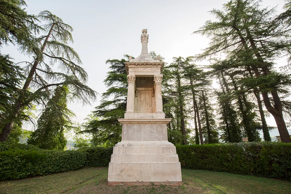 A white stone funerary monument on a stepped base in a lawn, with an open columned upper storey and a pointed top, framed by tall cedars and a low hedge.