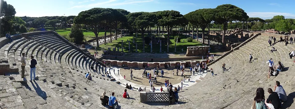 A wide panorama of a semicircle of stone seating stepping down to a flat orchestra, with visitors sitting on the steps and umbrella pines and columns beyond the stage.