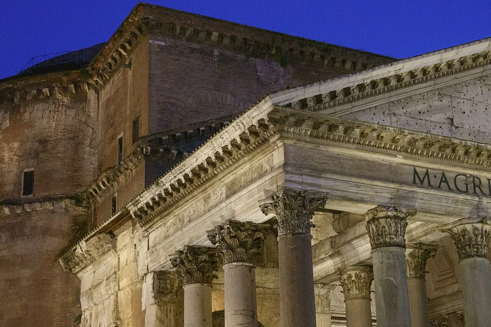 The portico of the Pantheon lit at dusk, Corinthian columns carrying a pediment and a lettered frieze, with the brick drum of the rotunda rising behind.