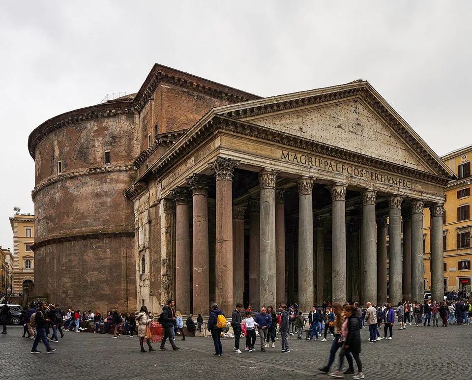 The Pantheon's portico of tall grey granite columns under a triangular pediment with a lettered frieze, the round brick drum of the rotunda rising behind it on the left, and a crowd on the paved square in front.