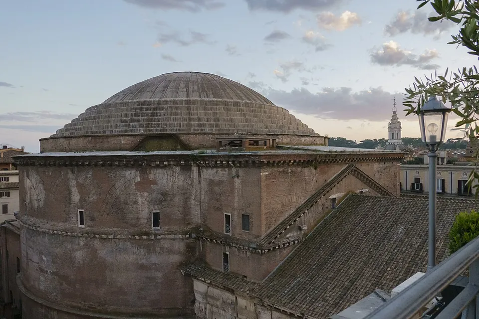 The Pantheon's low stepped dome and the plain brick drum beneath it, seen across rooftops at dusk, with a tower on the skyline to the right.