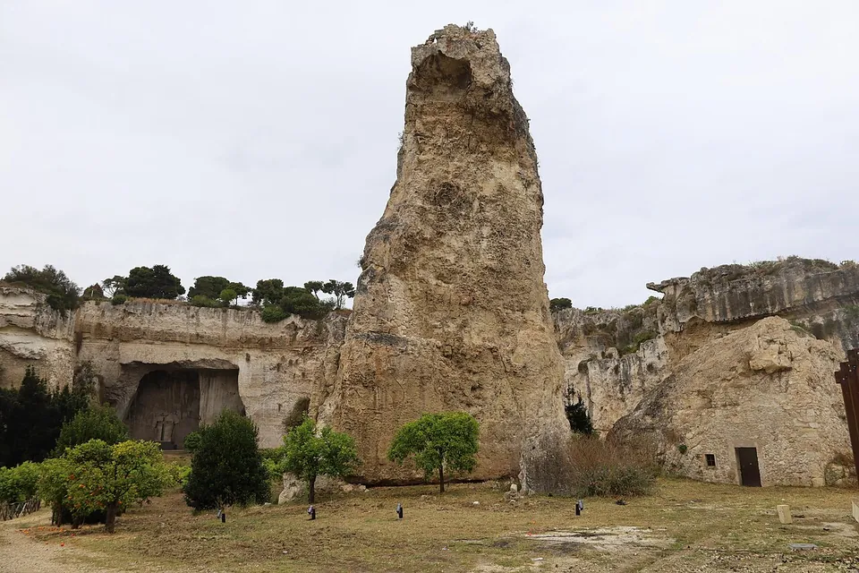 A tall, rough pillar of rock standing alone on the floor of a quarry, with cave openings in the cliff walls behind and young trees around its base.