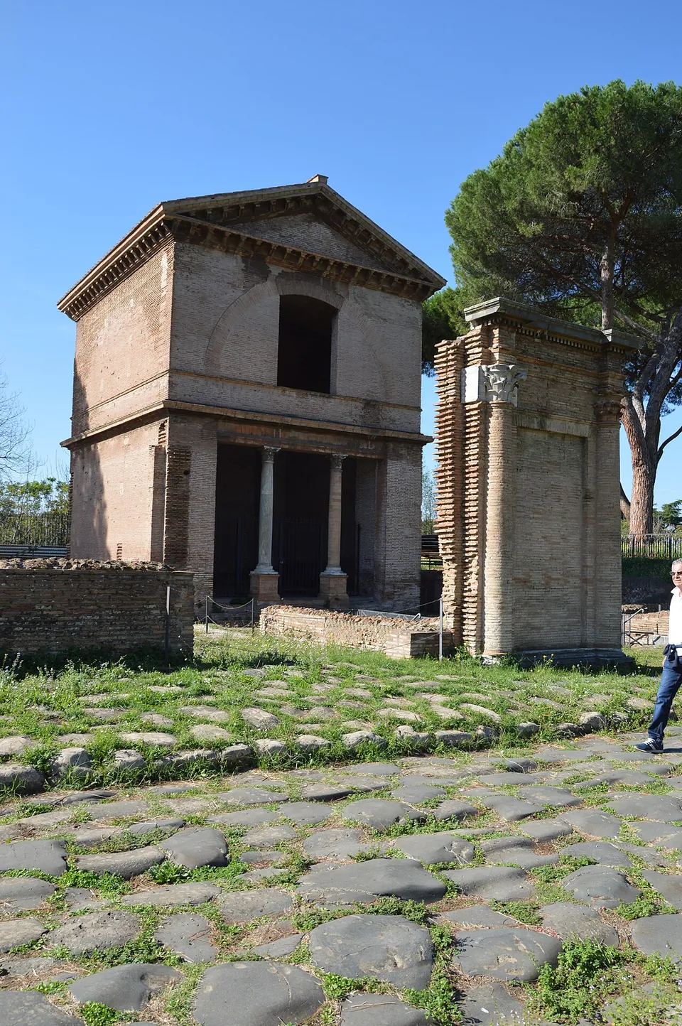 Large dark basalt paving stones of the Roman road in the foreground; beyond, a two-storey brick tomb with a pediment and two columns across its front, and beside it a tall, narrow brick pier.