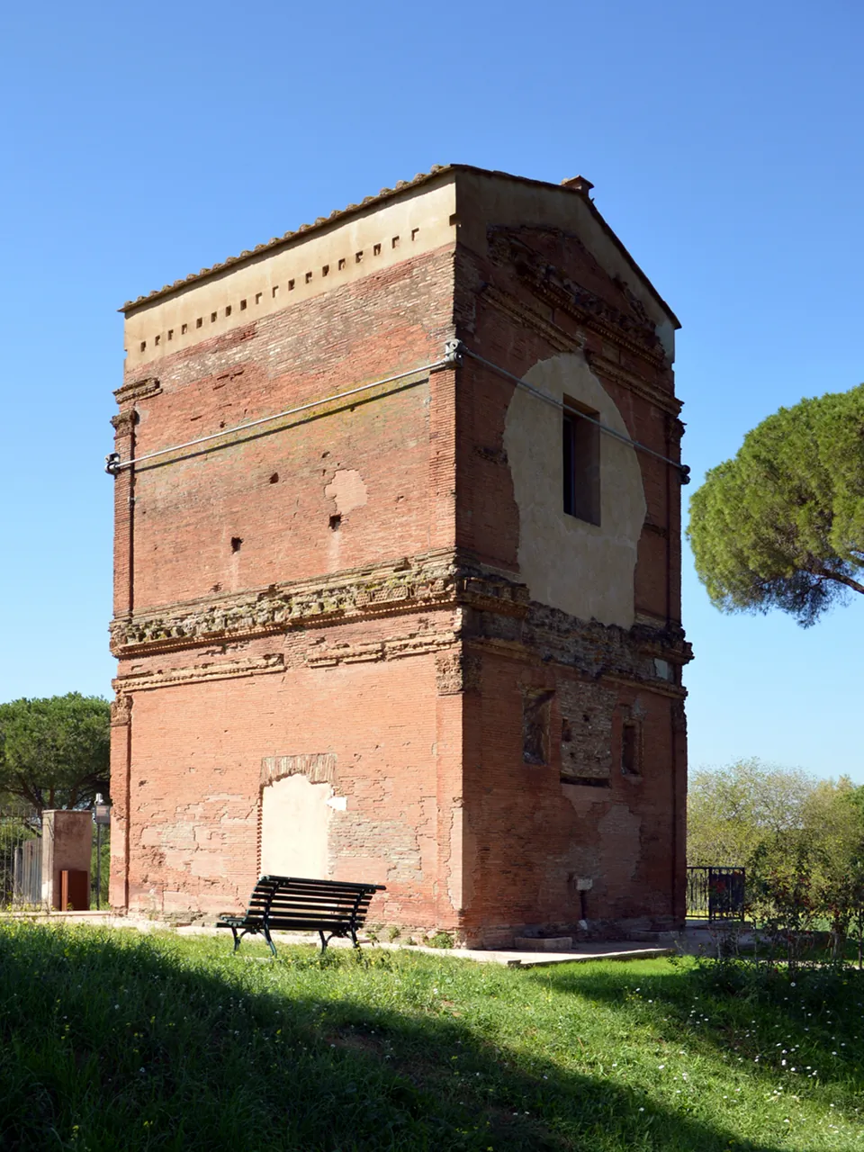 A tall, square tower of red and yellow brick with moulded cornices and a pitched roof, standing on a lawn with a bench in front of it.
