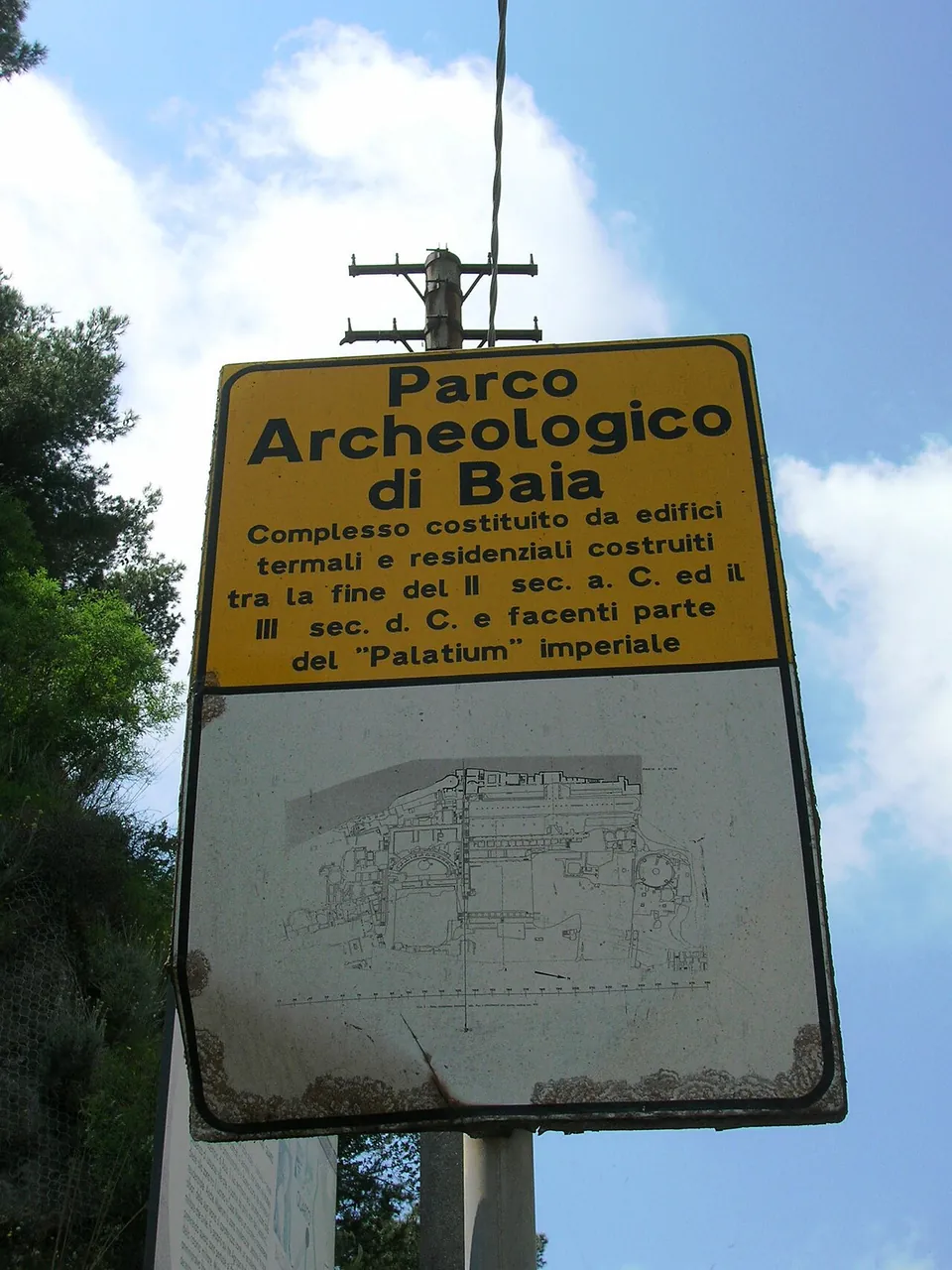 A yellow and white roadside sign reading "Parco Archeologico di Baia", describing a complex of bath and residential buildings built between the end of the 2nd century BC and the 3rd century AD as part of the imperial Palatium, above a plan of the site.