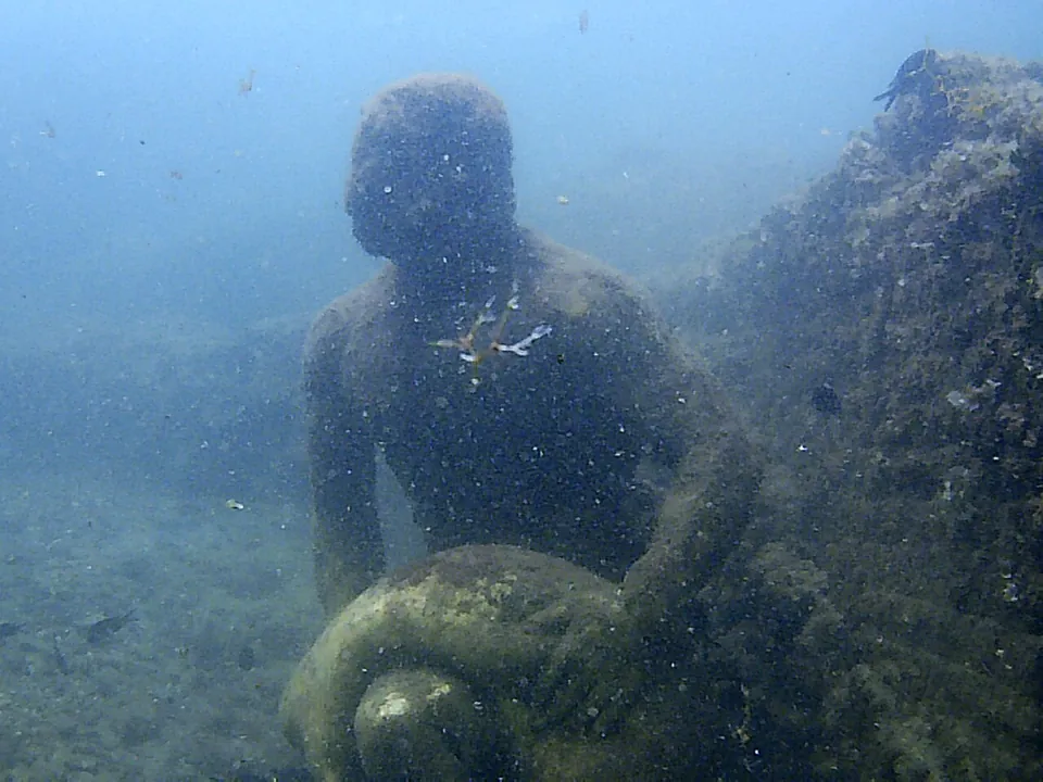 A statue of a figure bent over a large rounded object, encrusted with marine growth, on the sandy seabed in blue-green water.