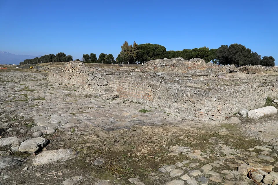 A cobbled surface in the foreground leading to a long, waist-high wall of stone set in a net-like pattern with bands of brick, a column drum lying at its end and umbrella pines behind.