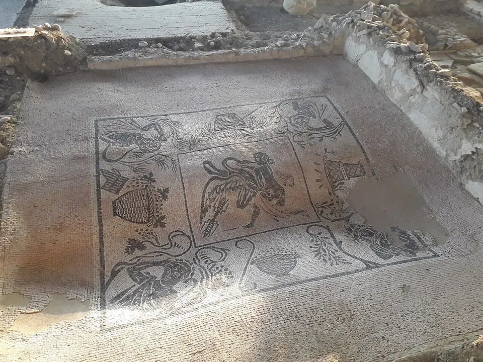 A black-and-white figured mosaic floor in a framed square, with a figure and a large bird in the central panel and baskets, vases and figures around it, partly stained and worn.