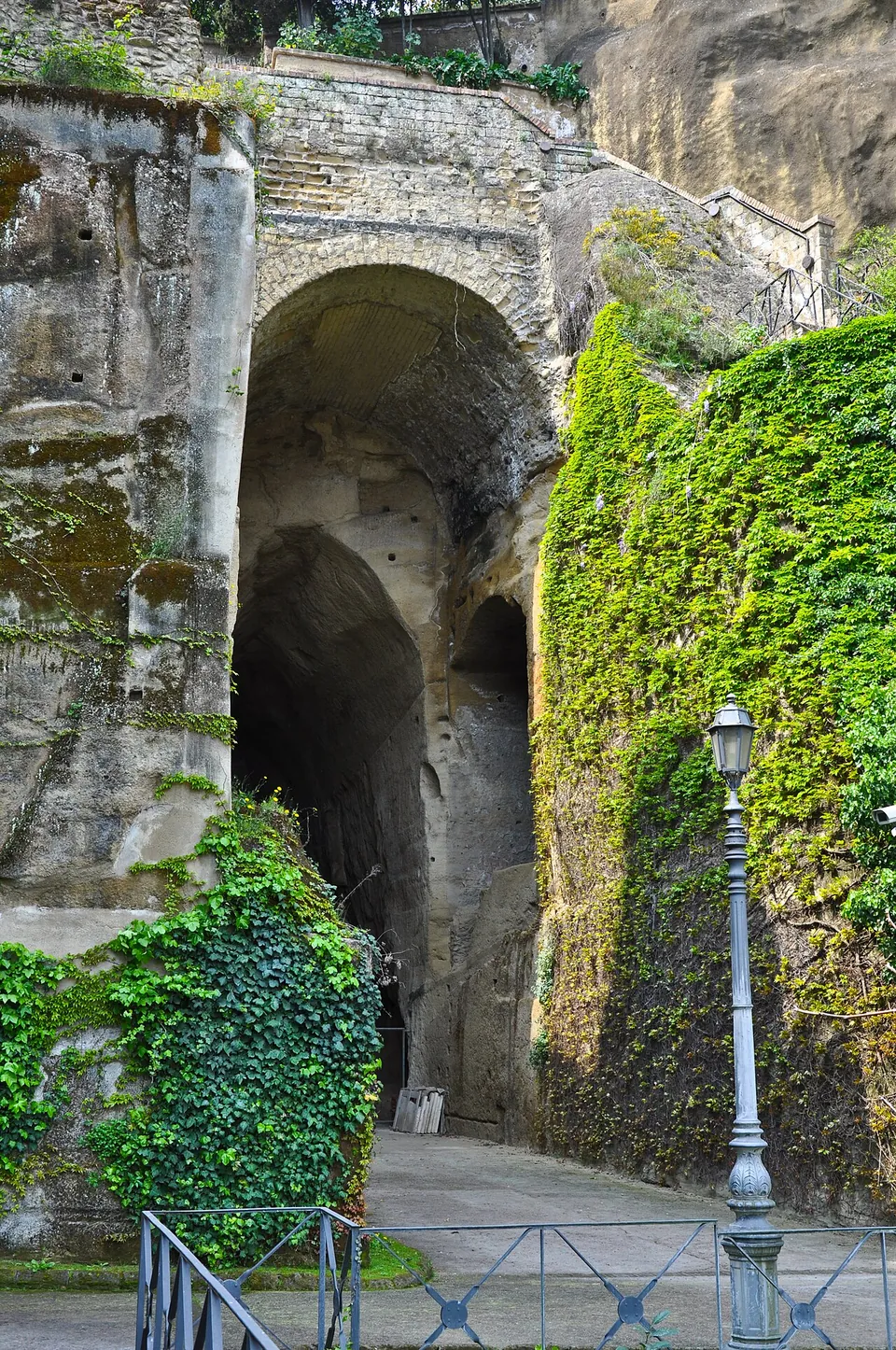 A tall arched opening in an ivy-hung rock face leads into a dark tunnel, with a paved path, a railing and a street lamp in front of it.