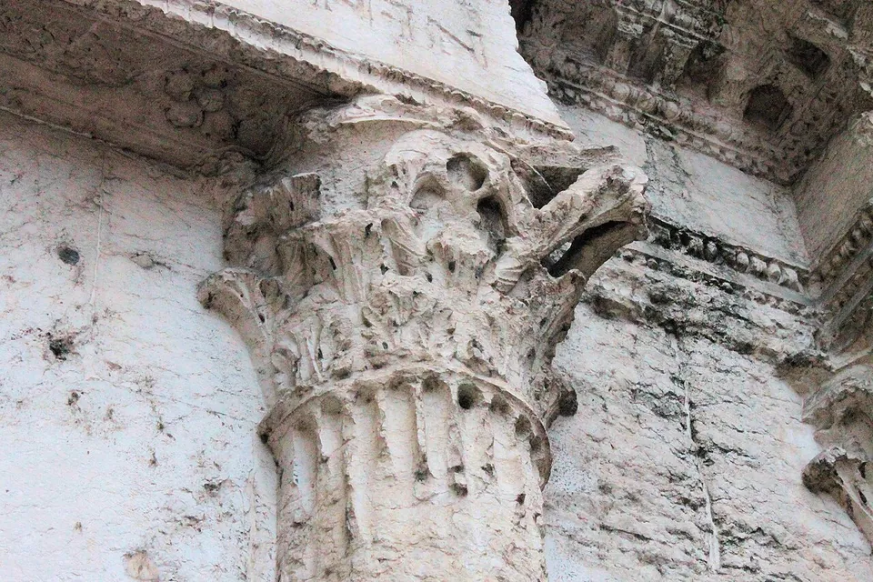 Close view of a weathered white stone capital carved with curling acanthus leaves, at the top of a fluted half-column set against the wall, under a carved cornice.