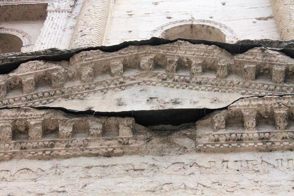 Looking up at the facade, a cracked horizontal cornice carved with rows of small blocks and beading, with strips of dark sheeting laid along its top and in the crack, a fluted column and an arched window above, and faint letters cut in the stone below.