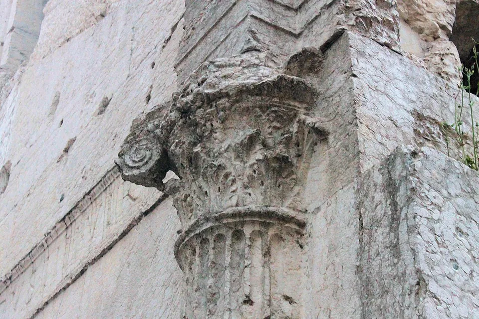 Close-up of a weathered capital of carved acanthus leaves on a fluted half-column, set against smooth blocks of pale stone.