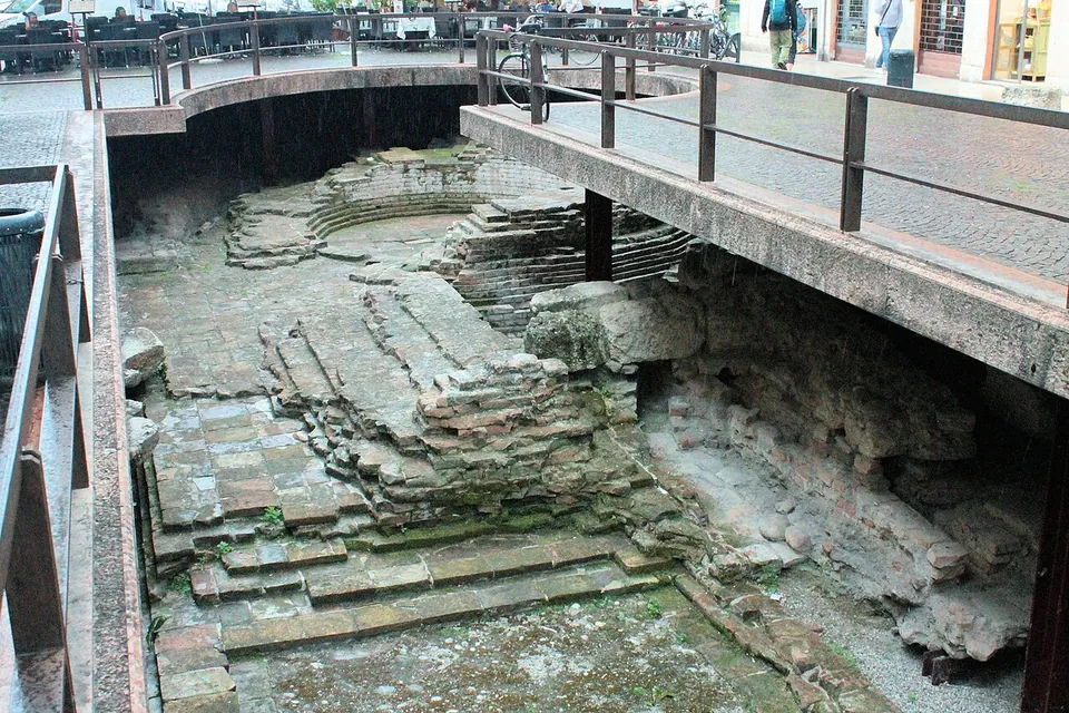 Looking down into a railed excavation below street level, where courses of stone and brick step down around a curved foundation, with the pavement, railings and café tables above.