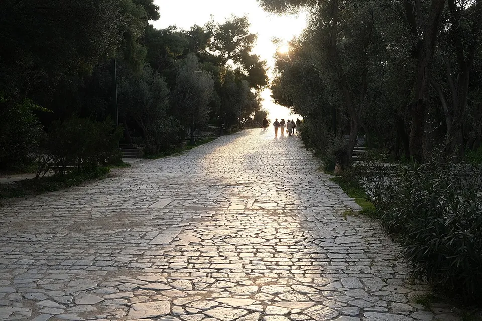 A broad path paved with irregular flat stones running between trees towards a low evening sun, with a few people walking far ahead.