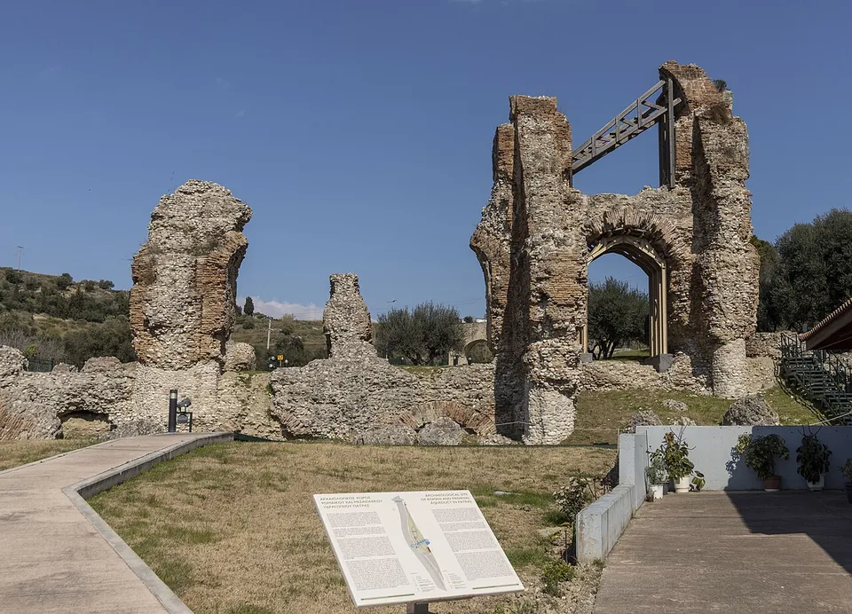 Tall ruined piers of rubble and brick, one still carrying an arch held by a modern steel brace, standing on a lawn with paved paths and an information board in front.