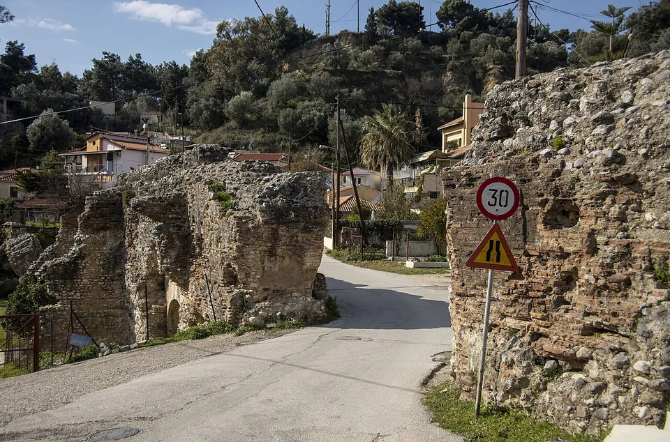 Massive ruined piers of rough stone on both sides of a narrow village road with traffic signs, houses and trees on the slope behind.