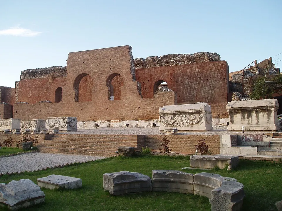 A tall brick wall with three arched openings rising behind a stage edged with carved marble blocks, and fragments of a curved marble parapet lying on the grass in front.