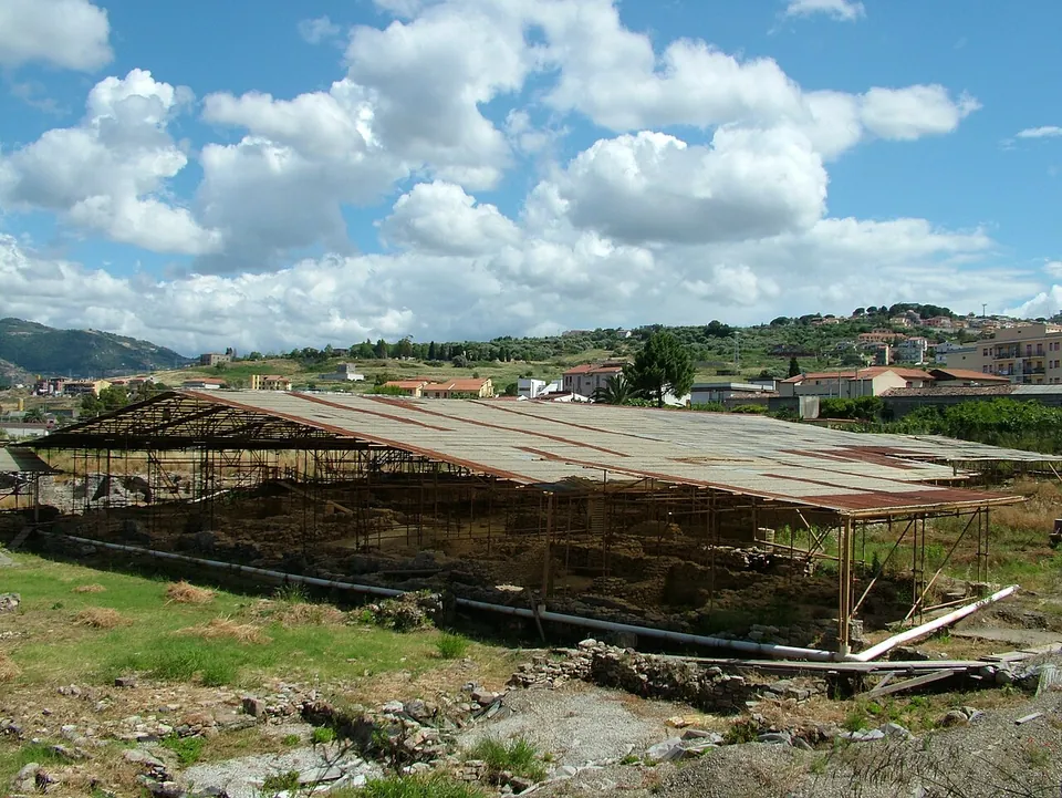 A long low roof of corrugated sheeting on a steel frame covering the excavations, with rough ground in front and the houses of Patti on the hillside behind under a cloudy sky.