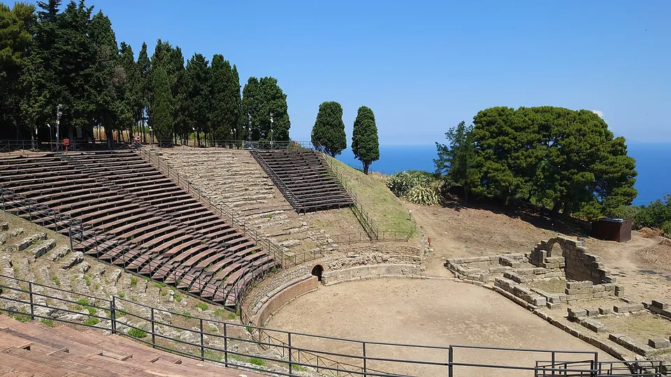The theatre seen across its floor, a flat sandy arena ringed by a low curved wall below the bottom row of seats, with the remains of a stone arch on the right and the sea beyond the trees.