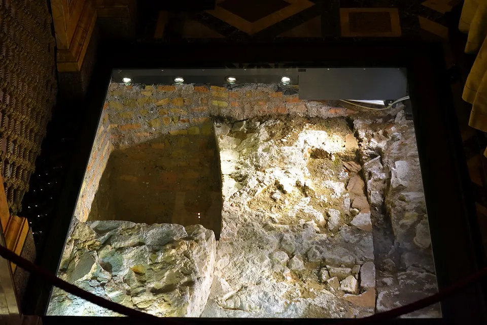 Looking down through a glass-covered opening in the floor onto rough stone and a short stretch of brick wall, lit from above, with a rope barrier around the edge.