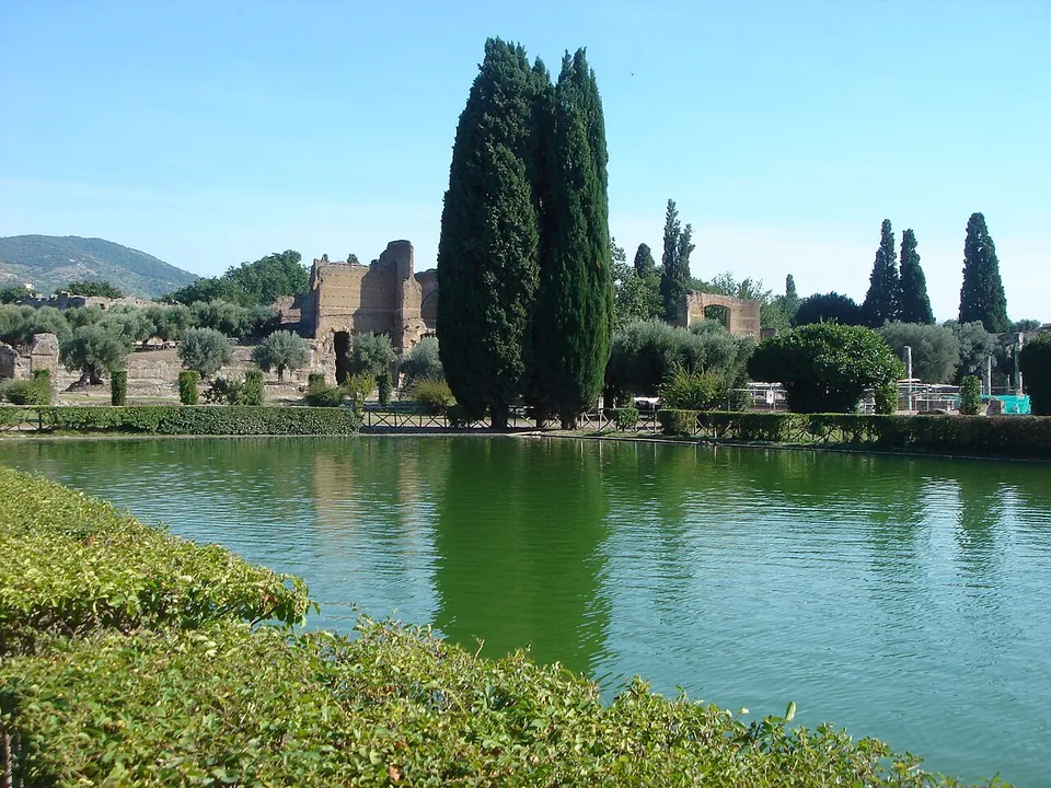 A broad pool reflecting tall cypresses and the ruined brick walls of buildings on its far side, with a clipped hedge in the foreground and hills on the horizon.