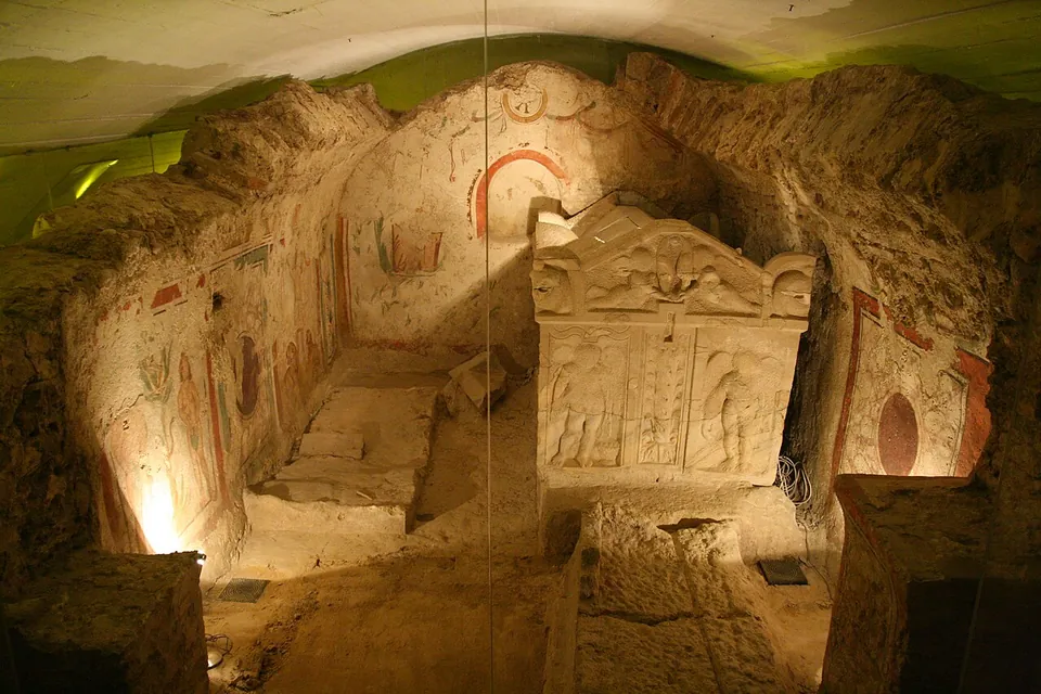 A lit underground burial chamber with walls painted in red, green and ochre figures and borders, a carved stone sarcophagus with figured panels standing in the middle.