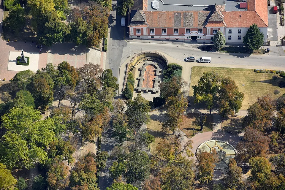 An aerial view of a park among trees and paths, with a sunken walled area of excavated foundations by a street and a long building with a tiled roof behind.