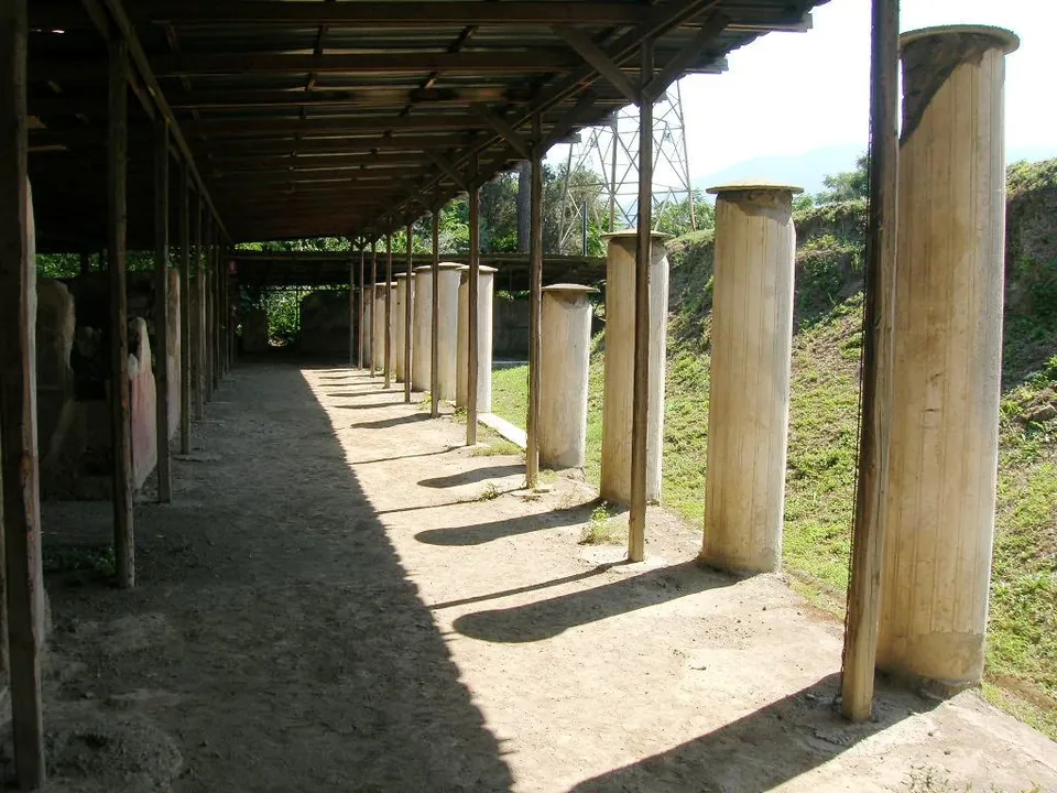 A row of plastered columns standing under a timber shelter roof beside a gravel walk, with a grassy bank on the right.