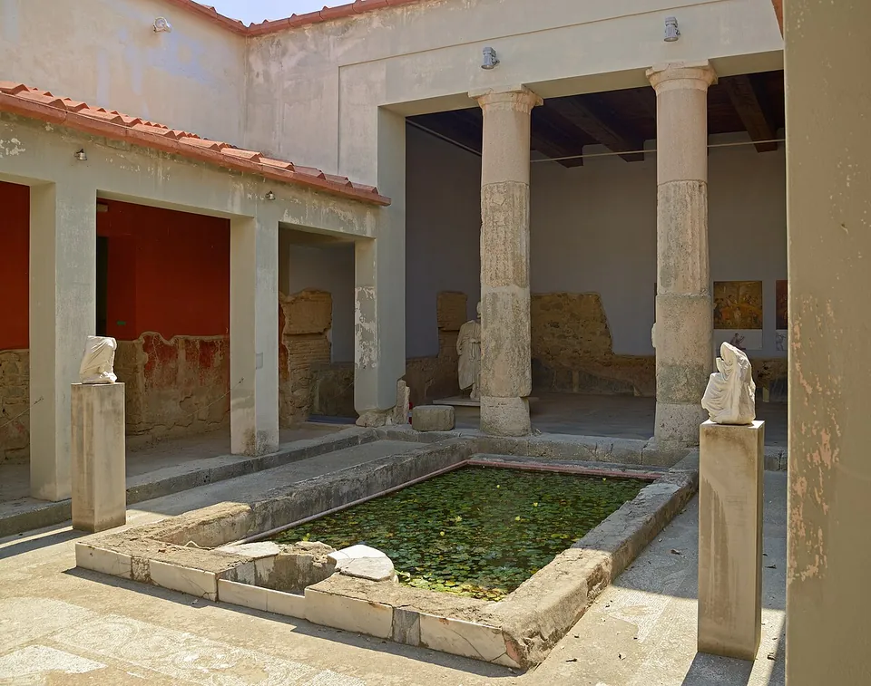 A small courtyard with a rectangular pool covered in water plants, two tall columns before a roofed room, red-painted walls and marble fragments on pedestals.