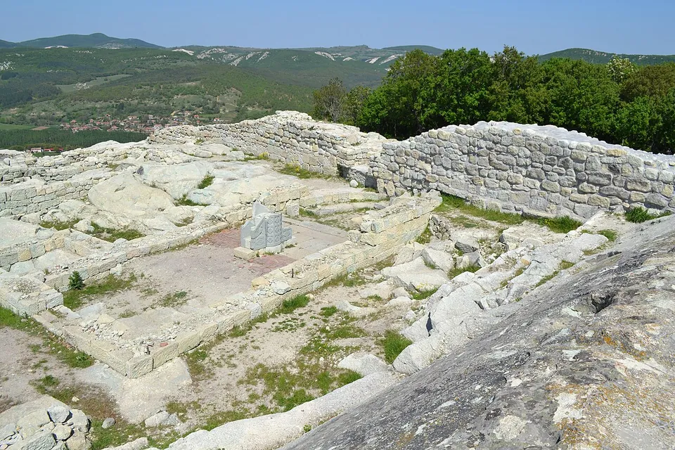 The excavated foundations of a rectangular building with a paved floor on the hilltop, edged by low stone walls and a higher stretch of wall, with green wooded hills and a village in the valley far below.