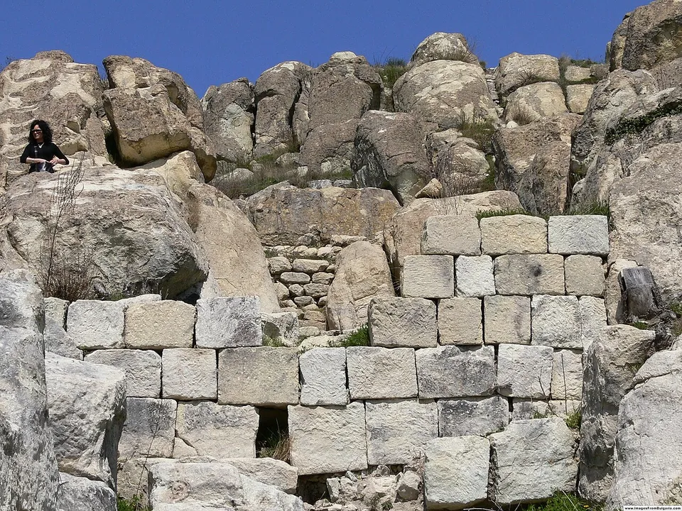 A wall of large squared stone blocks built up against weathered boulders of pale bedrock on a hilltop, with a visitor sitting on the rocks at the top left and a deep blue sky above.