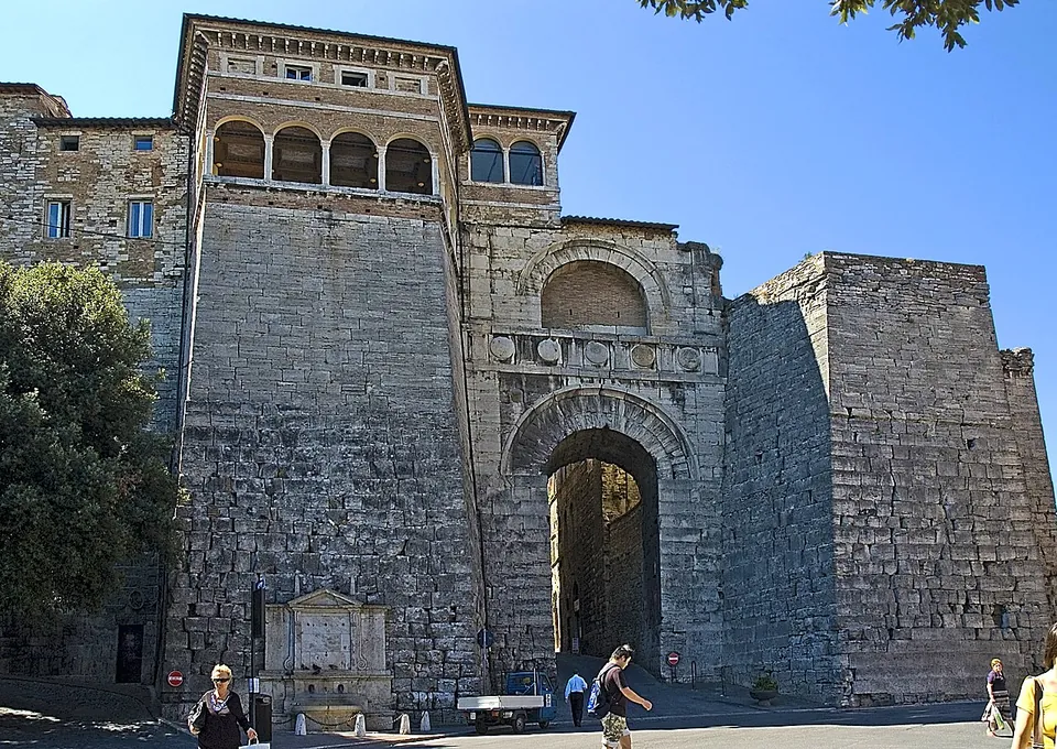 A massive gate of large pale travertine blocks seen from the square in front of it. Two tall sloping-sided towers flank a central front with a single round-headed archway at street level, a band of round shields above it and a second, smaller arch higher up. On top of the left tower stands a later storey with an open arcaded loggia, and a stone fountain is set against the foot of that tower. People walk past on the paving.