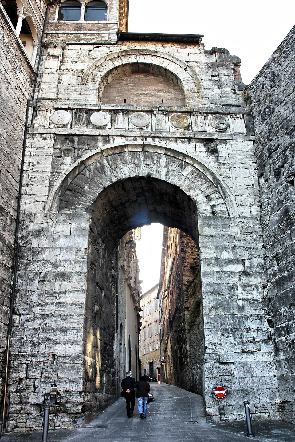 Looking straight into the archway of the gate from the street, its deep passage of big travertine blocks framing a narrow, rising street of old houses beyond, with two people walking through.