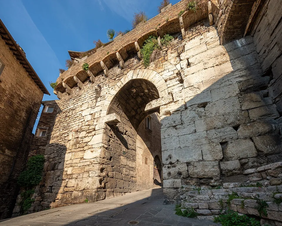 A gate of large, roughly squared pale stone blocks with a pointed arch, its upper edge lined with projecting stone corbels and tufts of plants, opening onto a narrow street in shadow.