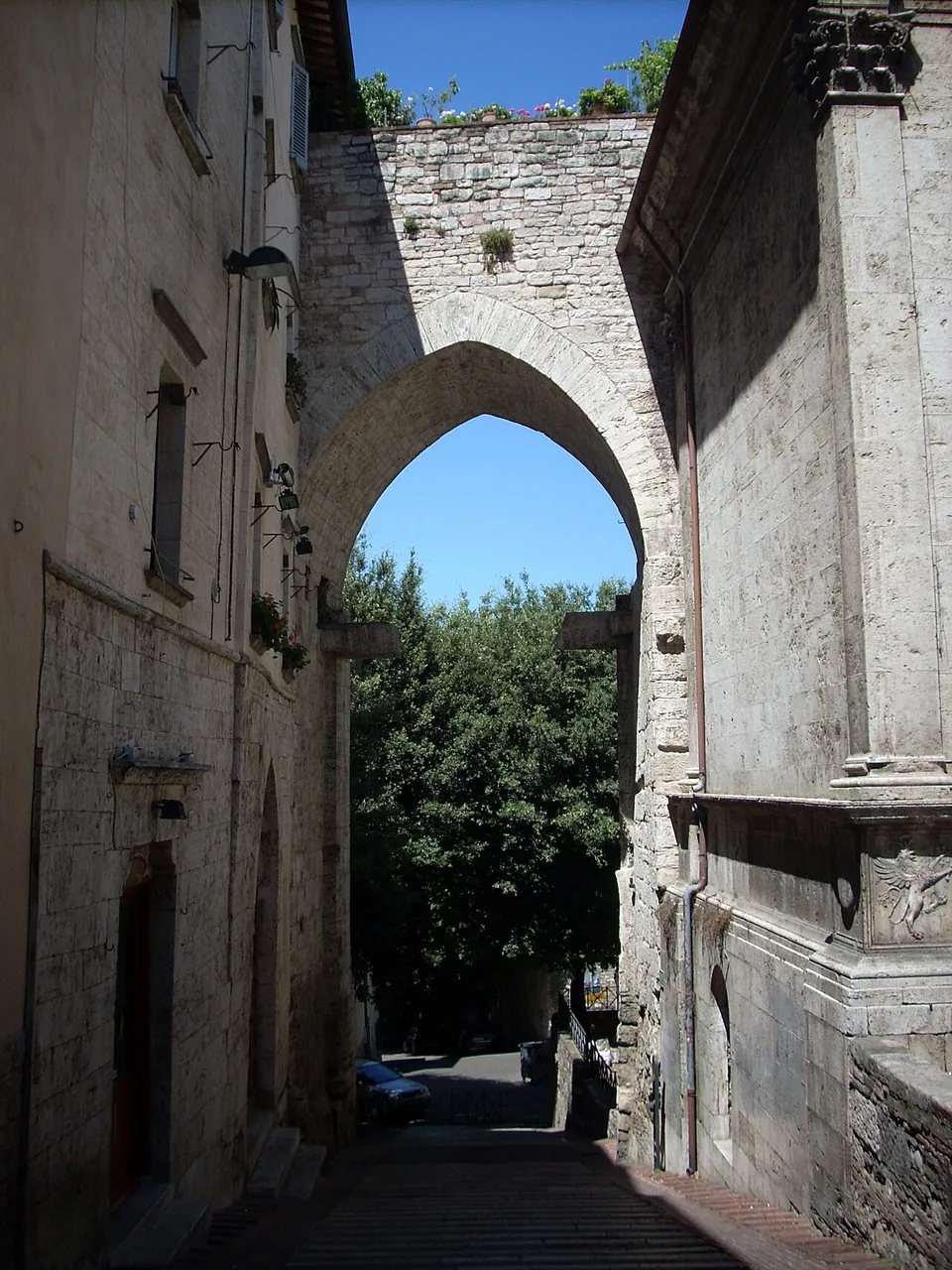 A high pointed archway in a stone wall at the end of a narrow lane between old buildings, with trees and sky showing through the opening.