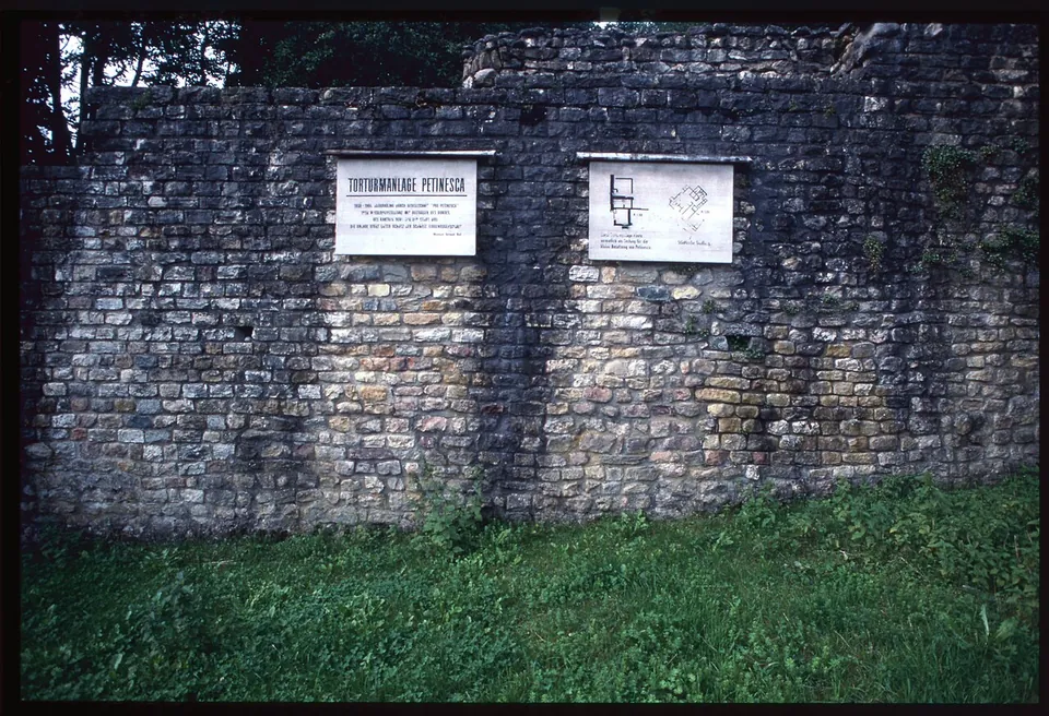 A long wall of small squared stones with two white plaques fixed to it, one headed "Torturmanlage Petinesca" and one showing a plan, above a strip of grass.