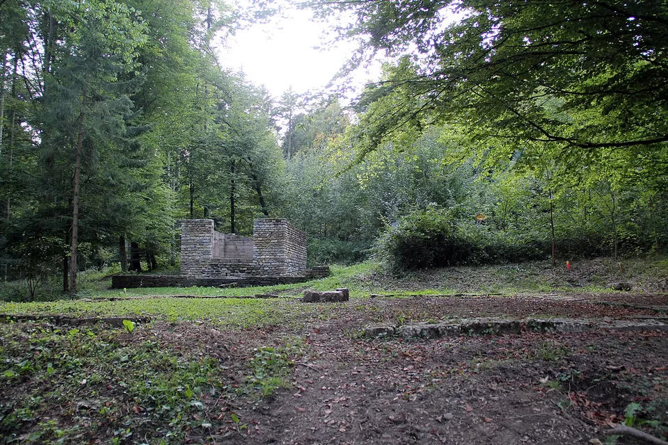 Low stone foundations in a clearing in the woods, with the squared walls of a small building standing waist to head high in the middle distance among tall trees.
