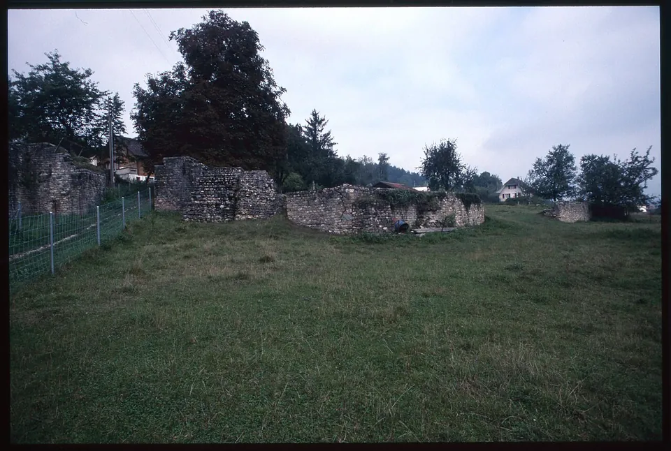 Ruined walls of rough stone standing in a mown meadow, a square block of masonry beside lower wall stubs, with trees and houses behind under a grey sky.