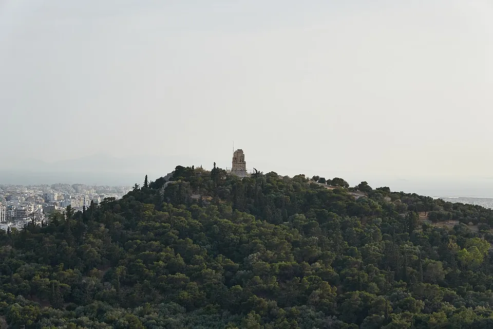 A rounded hill covered in dark green pines with a single pale stone monument on its summit, seen across the valley, with the city in the haze beyond.