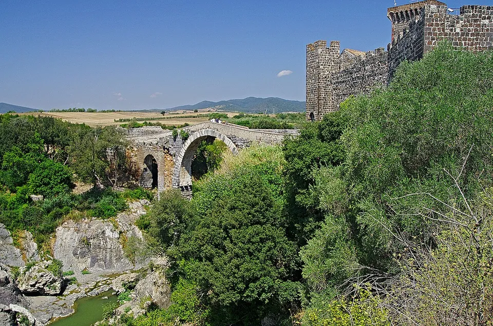 A wooded gorge with pale rocks and a green pool at the bottom; across the gorge a high stone arch, with a smaller one at its far end, carries the crossing to the dark battlemented walls of a castle, with open farmland and blue hills beyond.