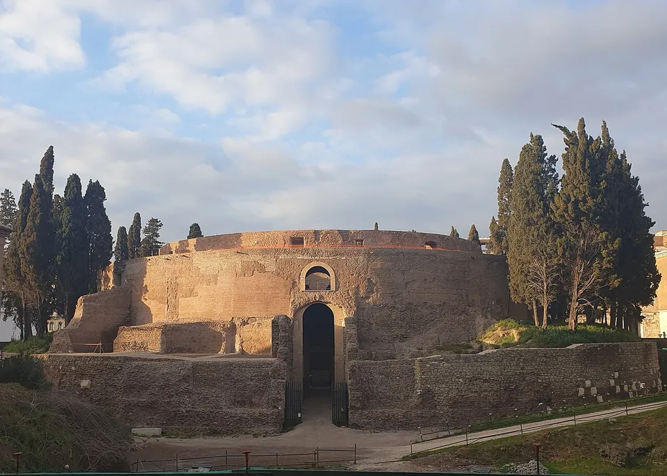 A broad, low round ruin of brick and grey stone under a cloudy sky, cypresses standing on and beside it, with a tall arched doorway in the middle of the front, a smaller arched window above it, and a sunken path leading up to the door between lower curved walls.