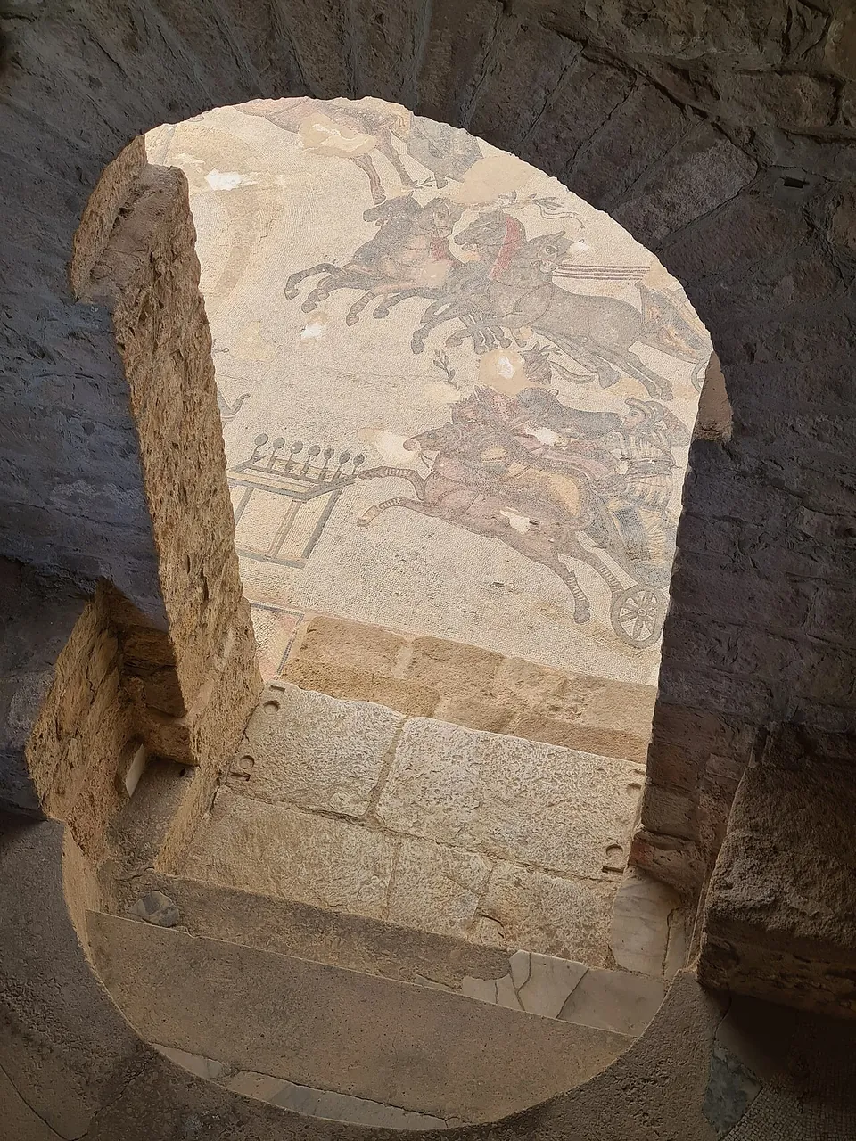 Seen through a narrow doorway of cut stone, part of a floor mosaic showing chariots drawn by galloping horses.