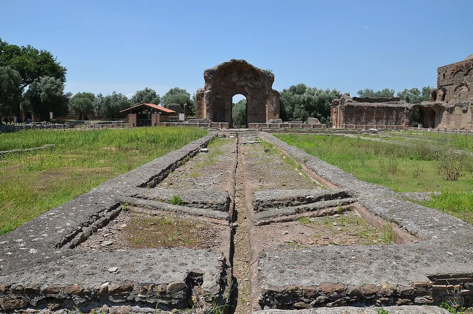 A long grassy courtyard with a channel of low stone basins running down its middle towards a tall ruined brick arch at the far end, with more ruined walls to the right and trees behind.