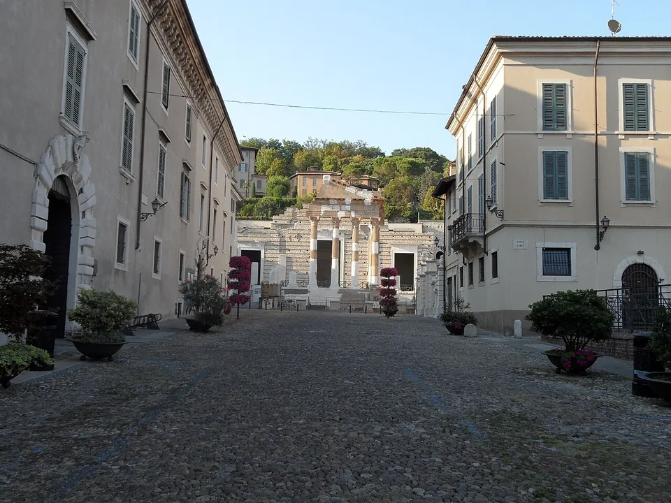 A cobbled square between pale three- and four-storey buildings with potted shrubs along their fronts, closed at the far end by the columned front of a ruined temple, with a wooded hill rising behind it.