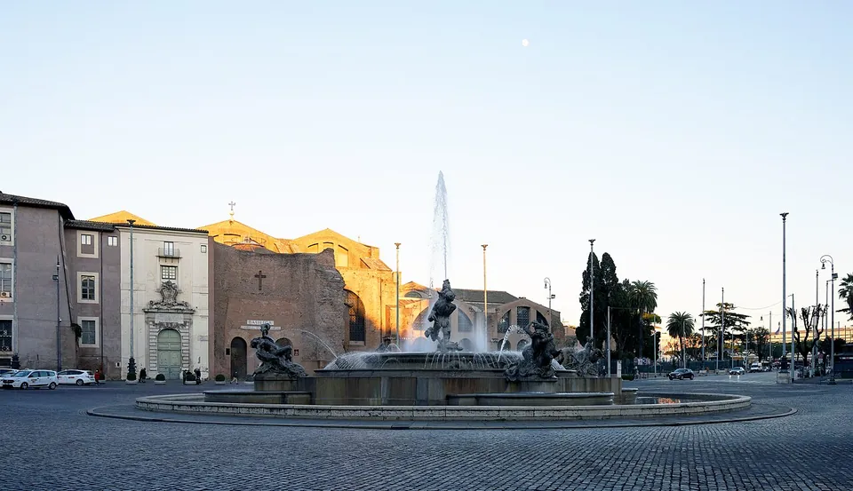 A wide paved square at dusk with a fountain throwing a tall jet in its middle, bronze figures round the basin, and at the far side a plain, unfaced brick building front between pale later houses.